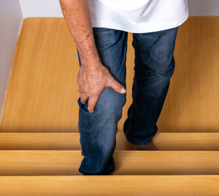 An elderly person holds their knee in pain while walking on wooden stairs, illustrating the challenges of a knee condition like a ligament injury or arthritis.