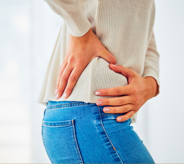  A woman in jeans and a sweater holds her hip with both hands, indicating pain or discomfort from an injury like a hip fracture.